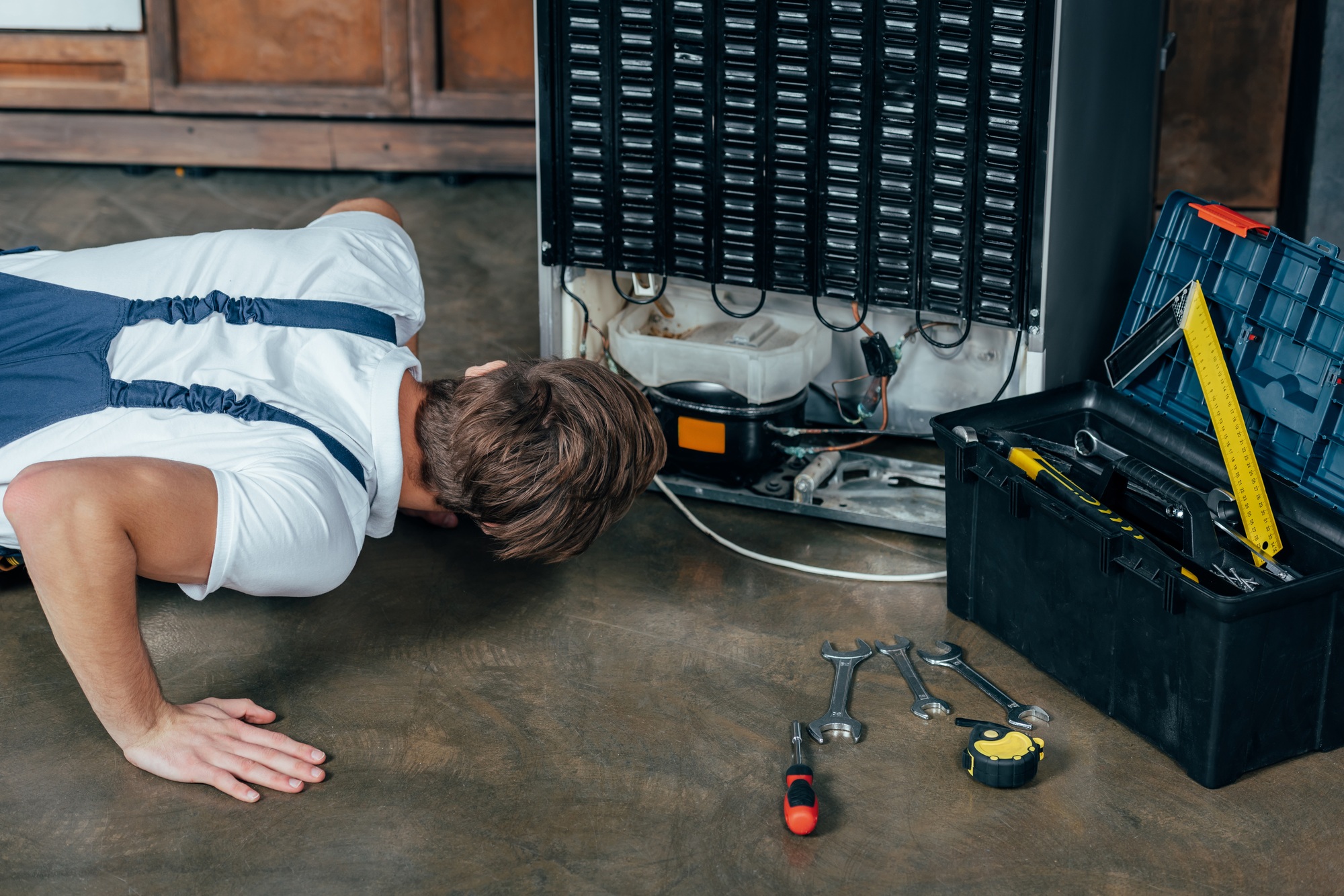 young repairman in protective workwear looking at broken refrigerator